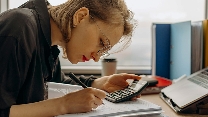 Woman working at desk, looking closely at what she's writing, holding a calculator in her left hand