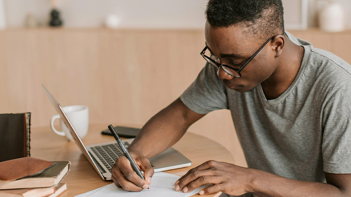 man writing on paper next to laptop